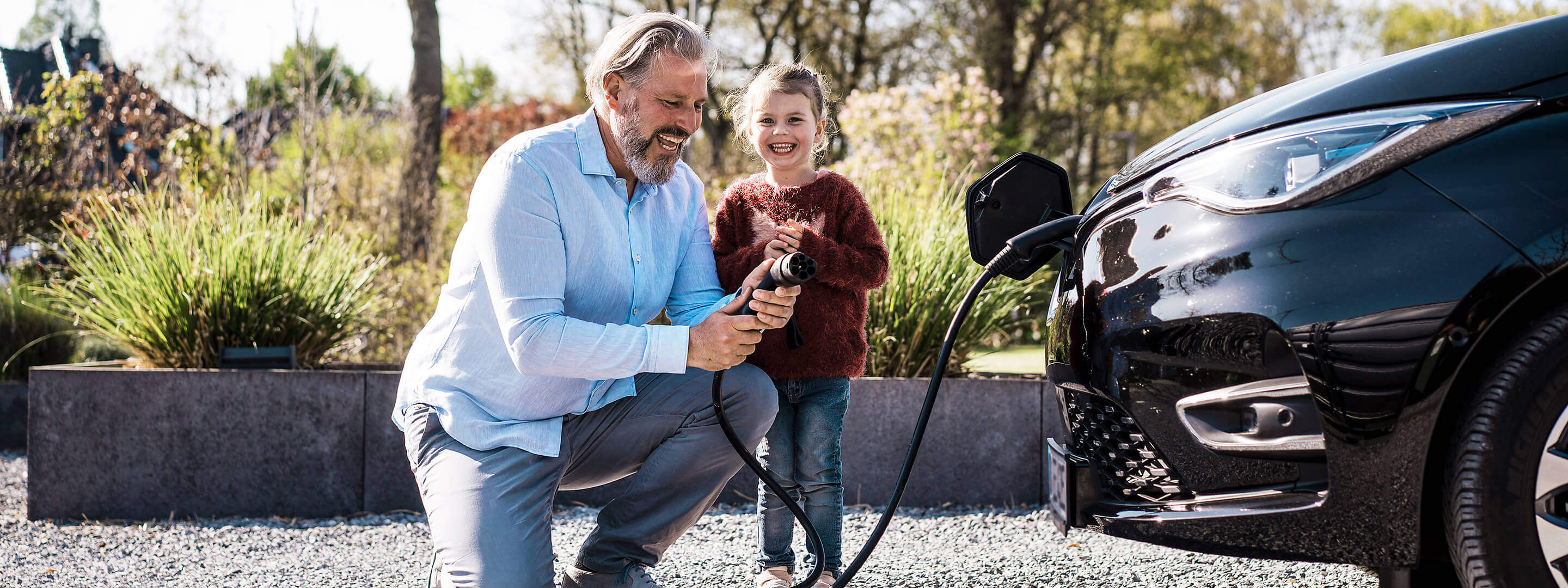 Mit Heim-Energie-Management-Systemen holst du mehr aus deinen Geräten raus Mann mit Tochter, der an einem sonnigen Tag vor einem E-Auto einen Ladekabel in der Hand hält