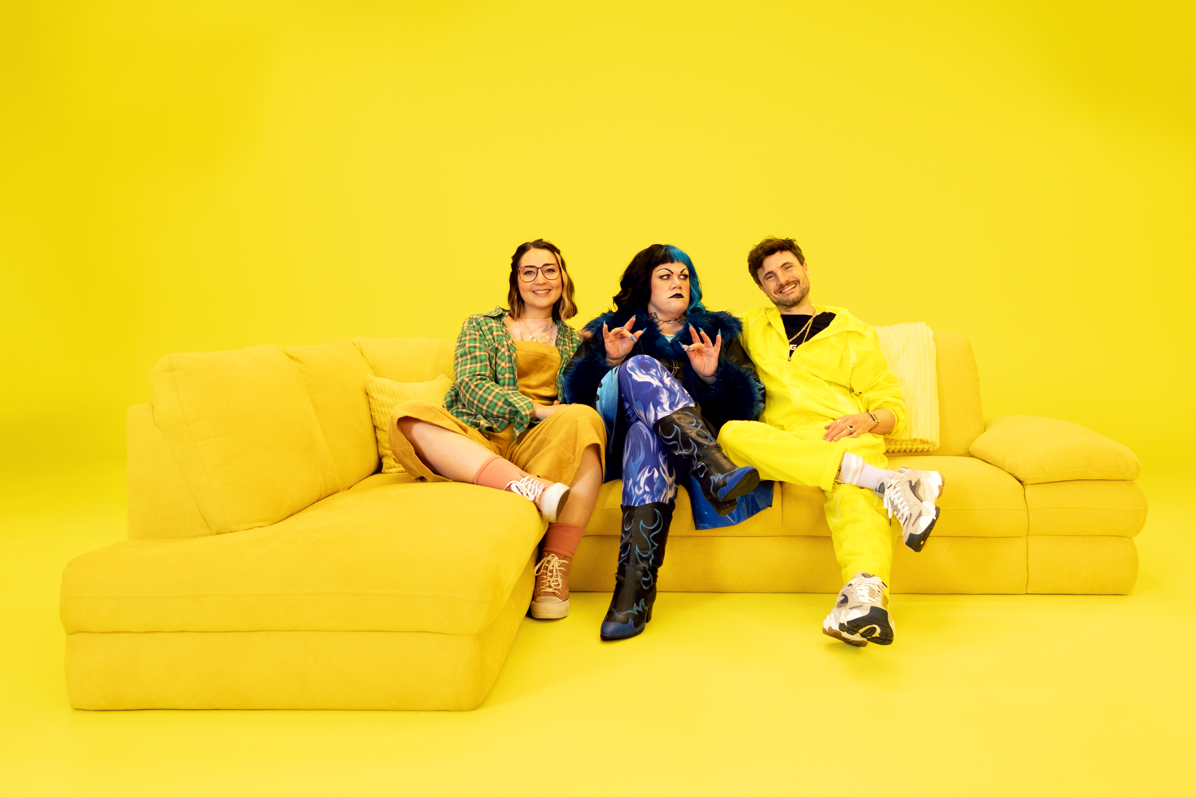 Drei Personen sitzen auf einem gelben Sofa vor gelbem Hintergrund, tragen bunte, modische Kleidung und lässige Schuhe.