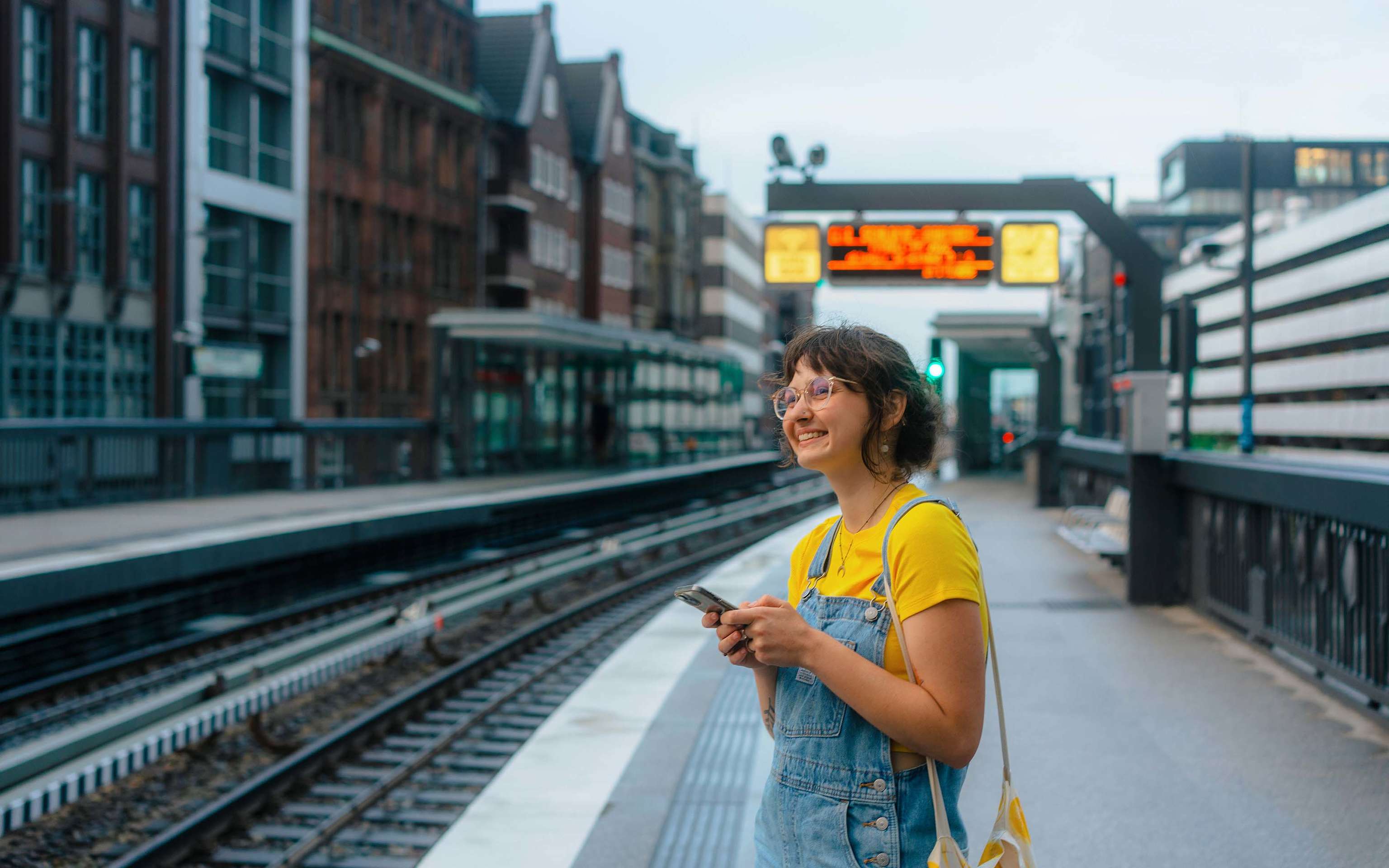 Eine junge Frau in einem gelben T-Shirt wartet mit ihrem Handy in der Hand auf eine Straßenbahn Eine junge Frau in einem gelben T-Shirt wartet mit ihrem Handy in der Hand auf eine Straßenbahn