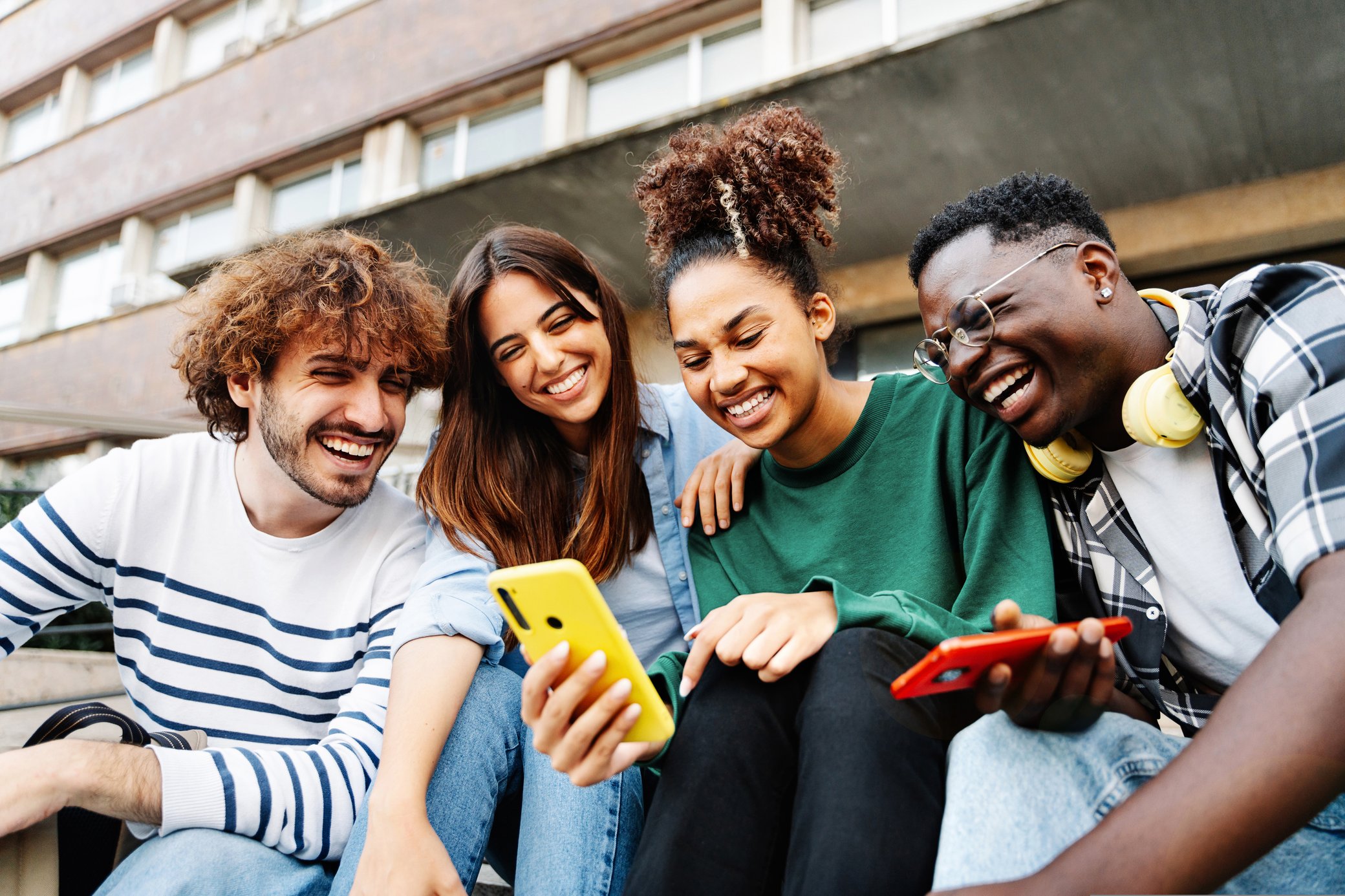 4 Freunde sitzen zusammen draußen vor einem Gebäude und schauen aufs Handy