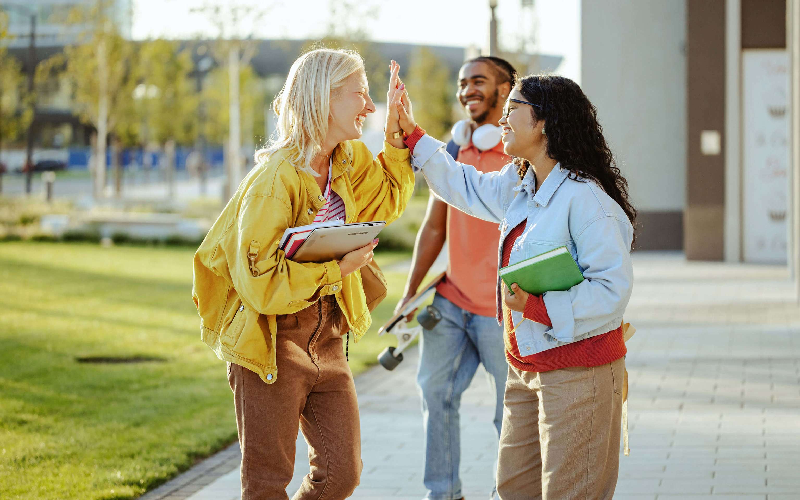 Zwei junge Frauen geben sich lachend ein High-Five, ein Freund mit Skateboard begleitet sie im Hintergrund Zwei junge Frauen geben sich lachend ein High-Five, ein Freund mit Skateboard begleitet sie im Hintergrund