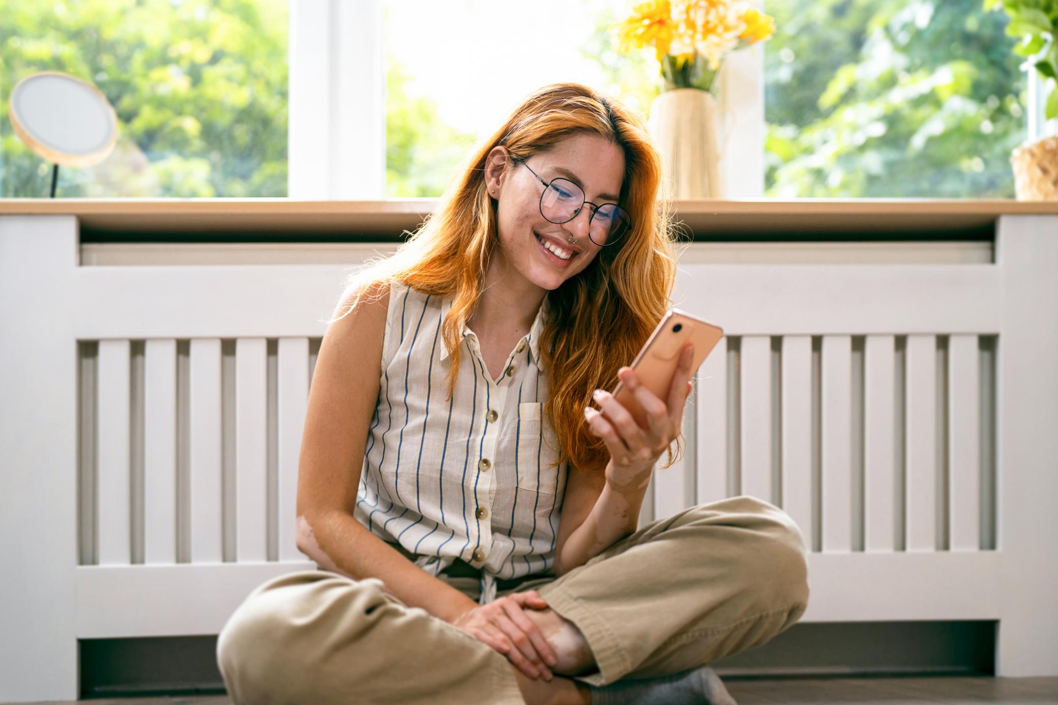 Frau sitzt vor ihrer Heizung und schaut  auf ihr Smartphone.
