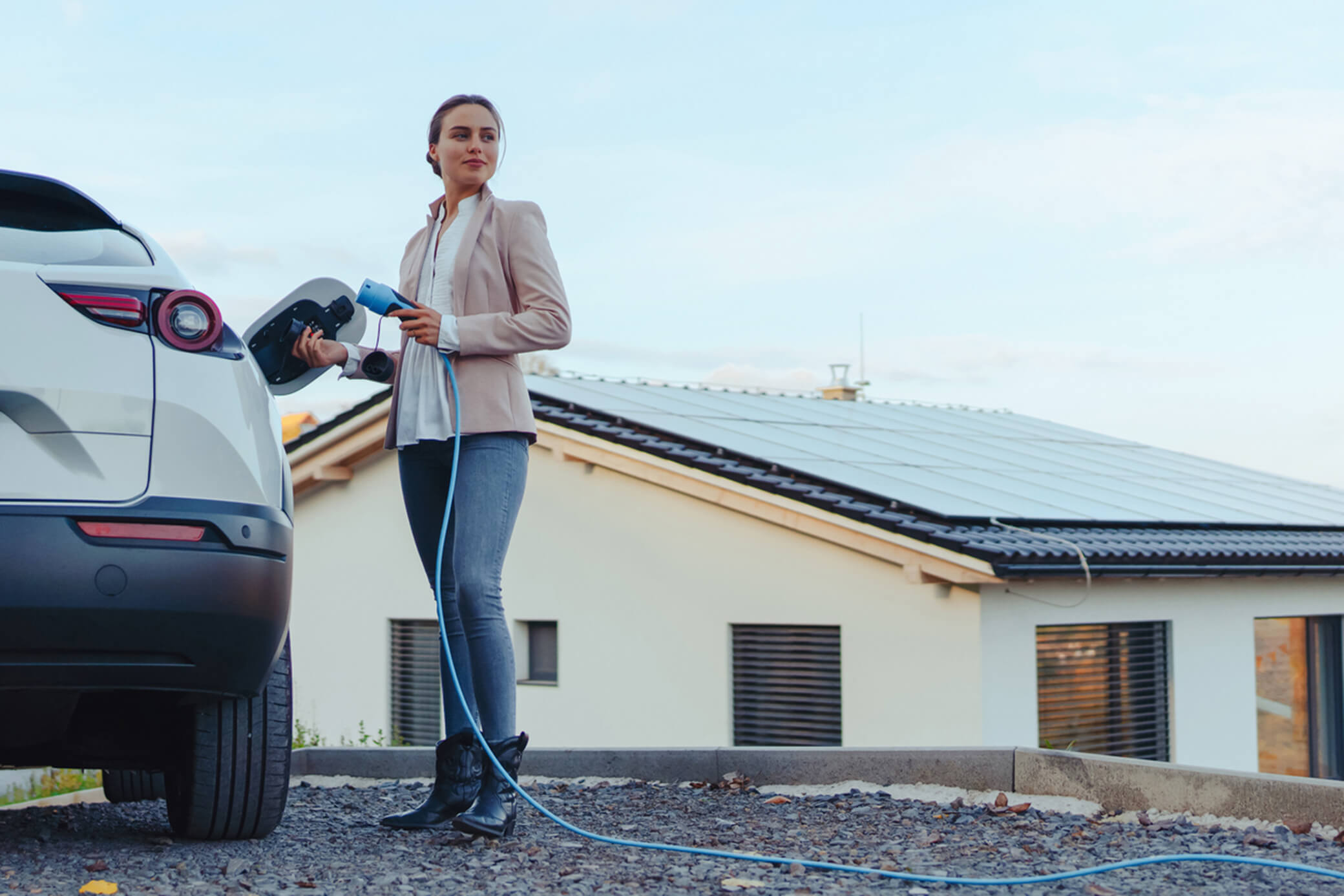Eine Frau lädt ein weißes Elektroauto und hält ein Ladekabel in der Hand - im Hintergrund ist eine Solaranlage