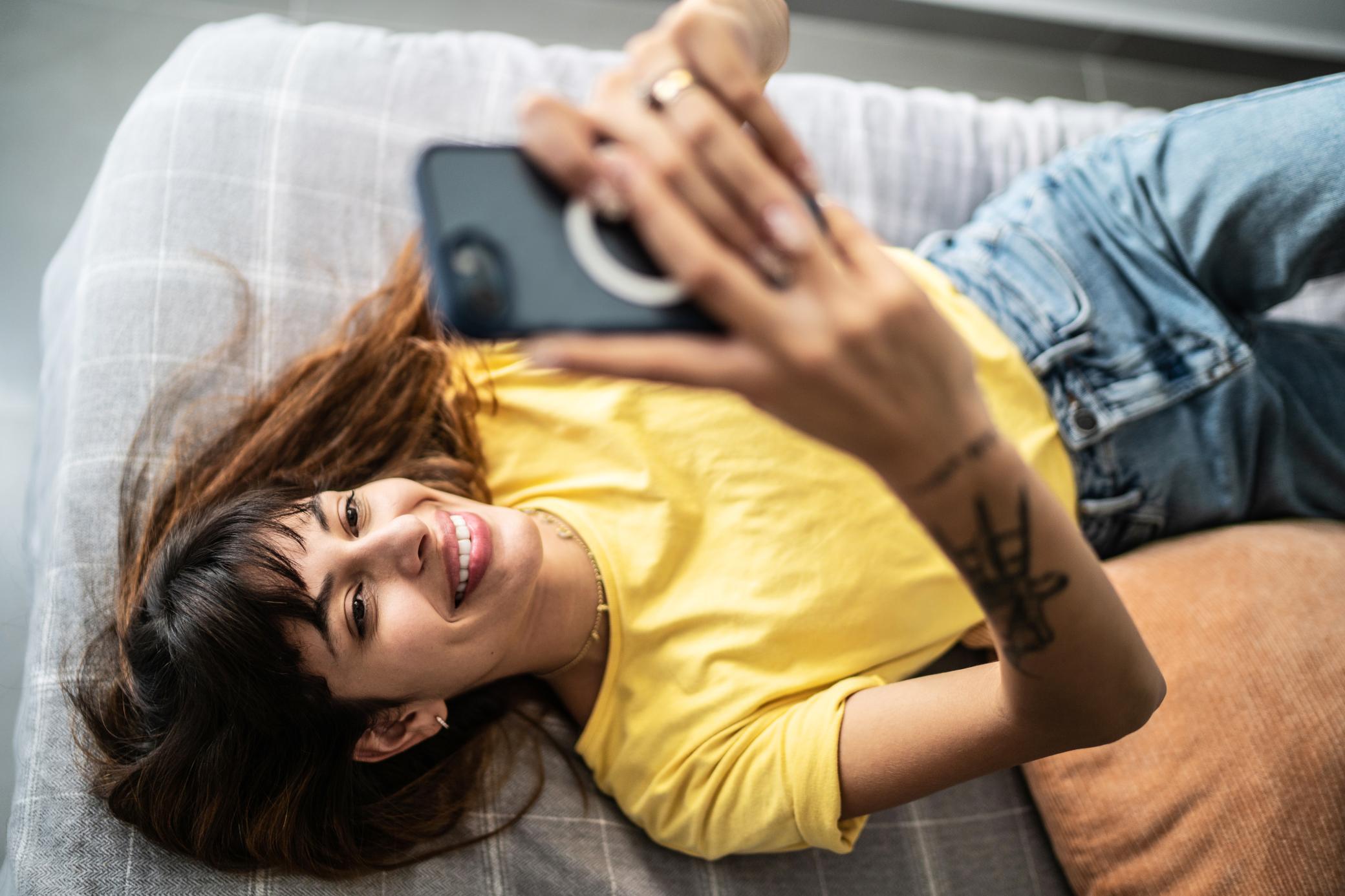 Eine junge Frau im gelben T-Shirt liegt auf dem Sofa und schaut aufs Handy