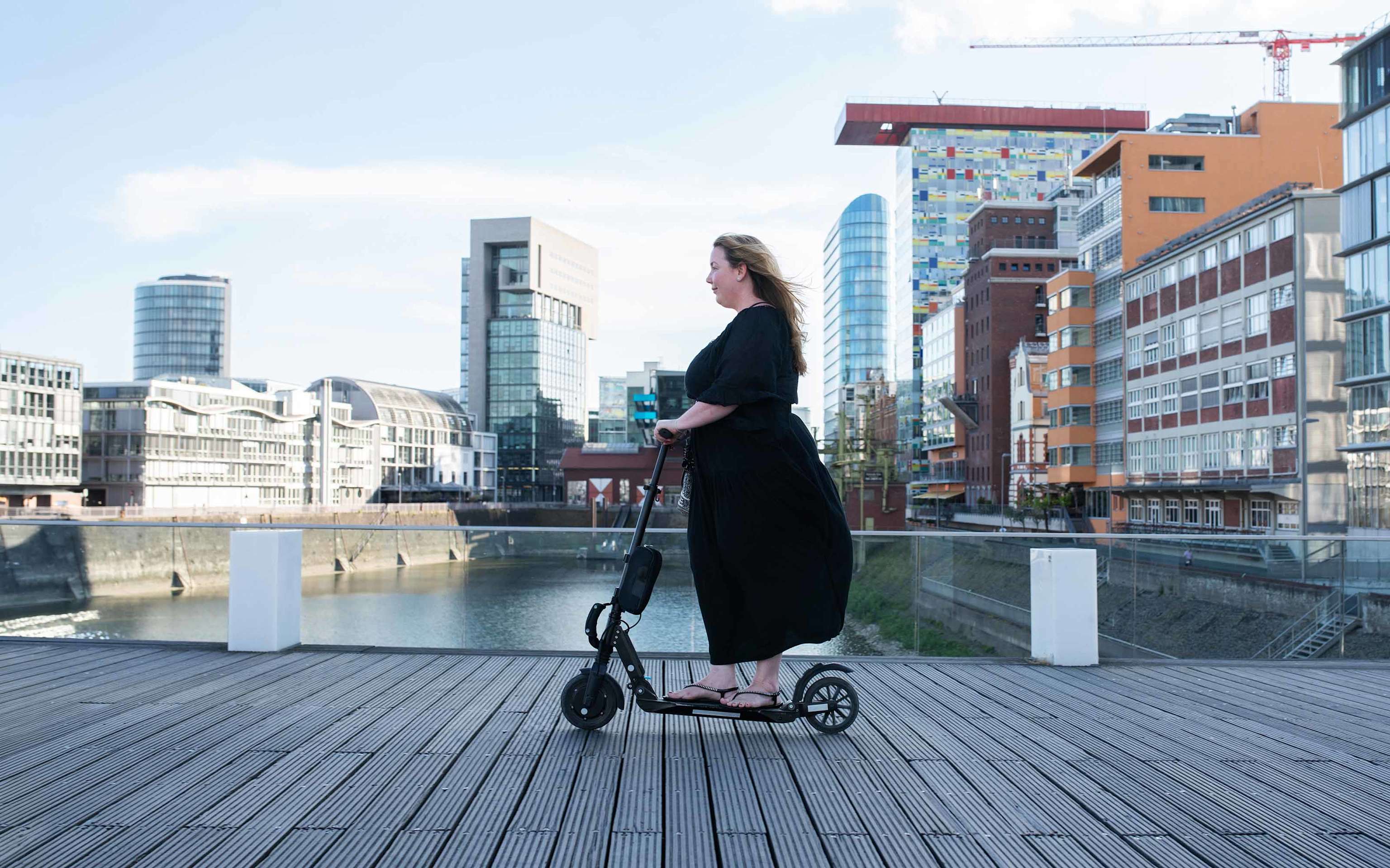 Frau mit schwarzem Kleid rollt auf E-Scooter durch den Rheinhafen in Düsseldorf Frau mit schwarzem Kleid rollt auf E-Scooter durch den Rheinhafen in Düsseldorf