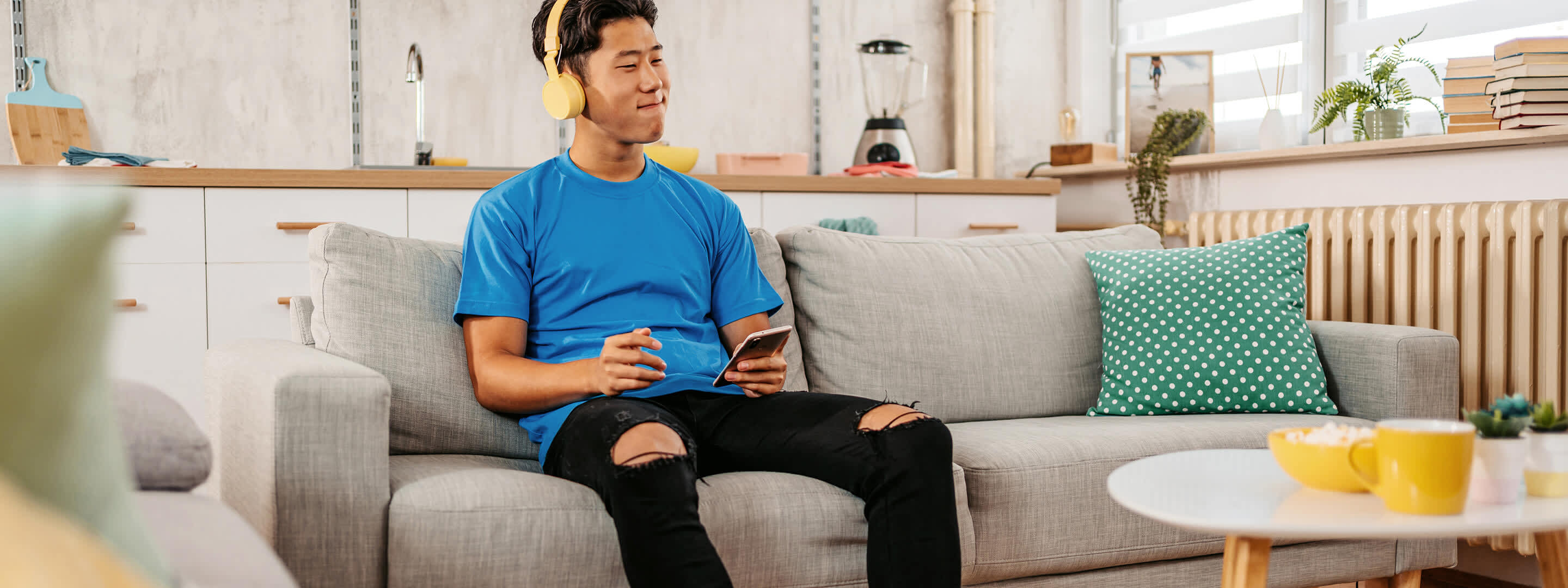 Junger Mann im blauen T-Shirt und gelben Kopfhörern sitzt auf der Couch und hört Musik Junger Mann im blauen T-Shirt und gelben Kopfhörern sitzt auf der Couch und hört Musik