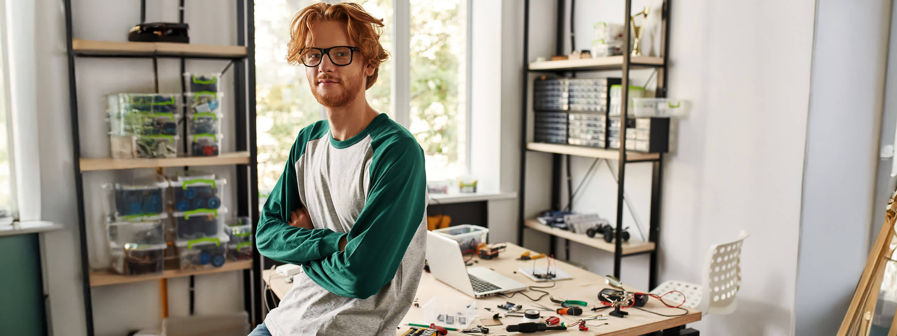 Mann sitzt auf dem Tisch im Home-Office - im Hintergrund sind elektrische Bauteile zu sehen Mann sitzt auf dem Tisch im Home-Office - im Hintergrund sind elektrische Bauteile zu sehen