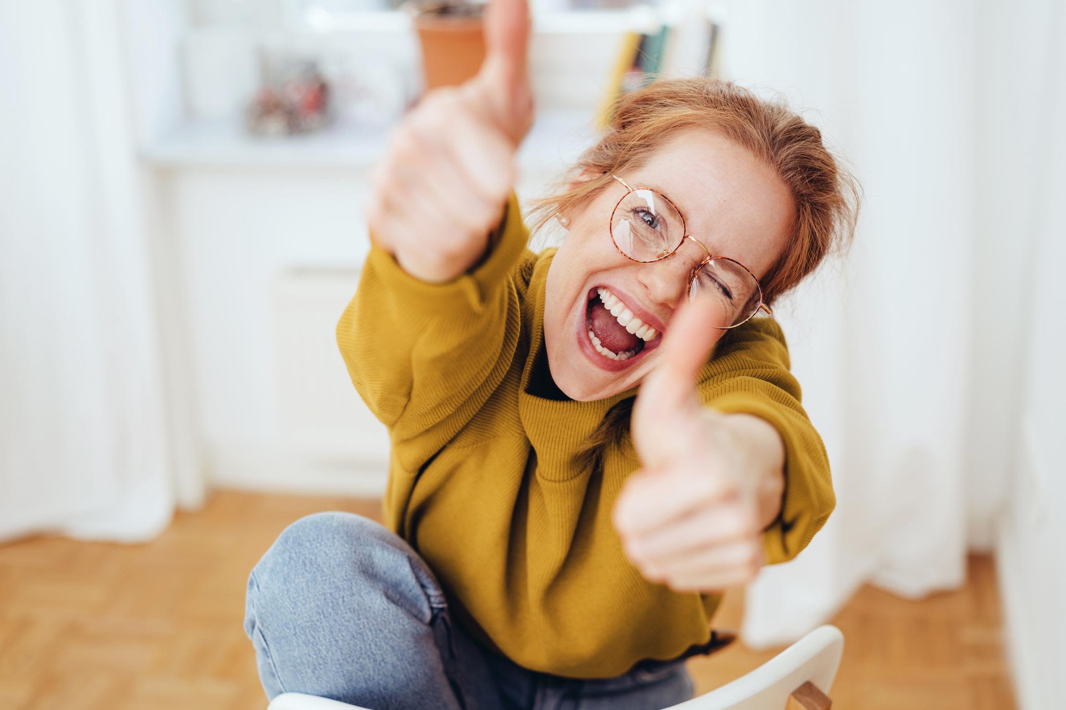 Kennst du deinen Zählerstand, hast du auch deinen Stromabschlag im Griff Frau mit Brille und gelbem Pulli zeigt happy beide Daumen hoch in ihrer Wohnung
