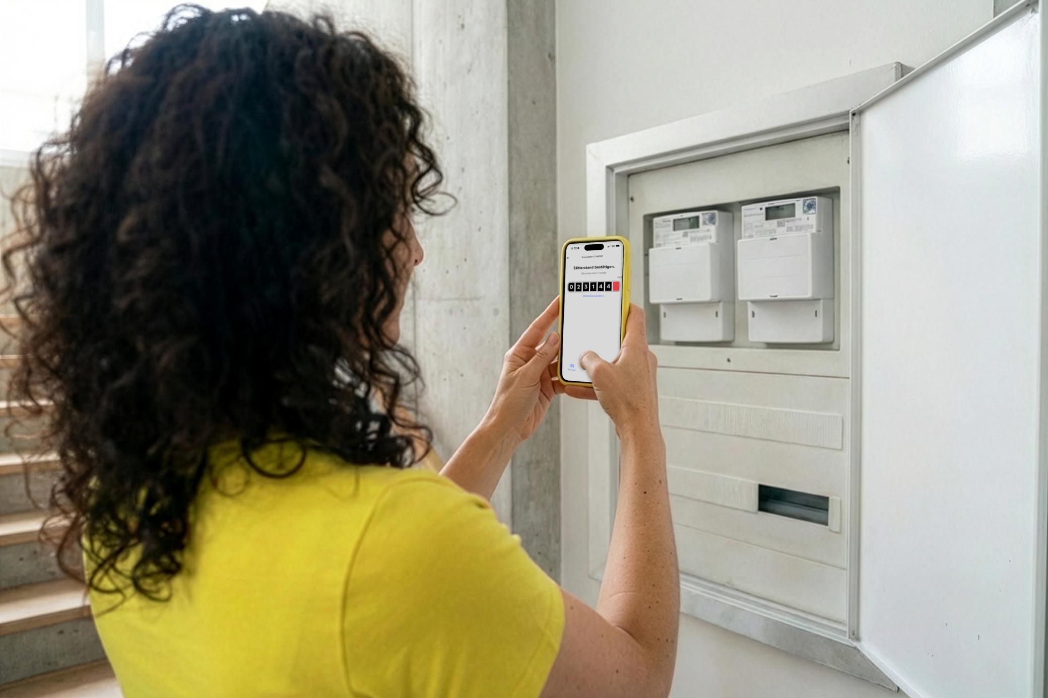 Frau mit gelbem Shirt fotografiert Zählerstand mit Handy und Yello App am Zählerkasten im Keller - Nahaufnahme