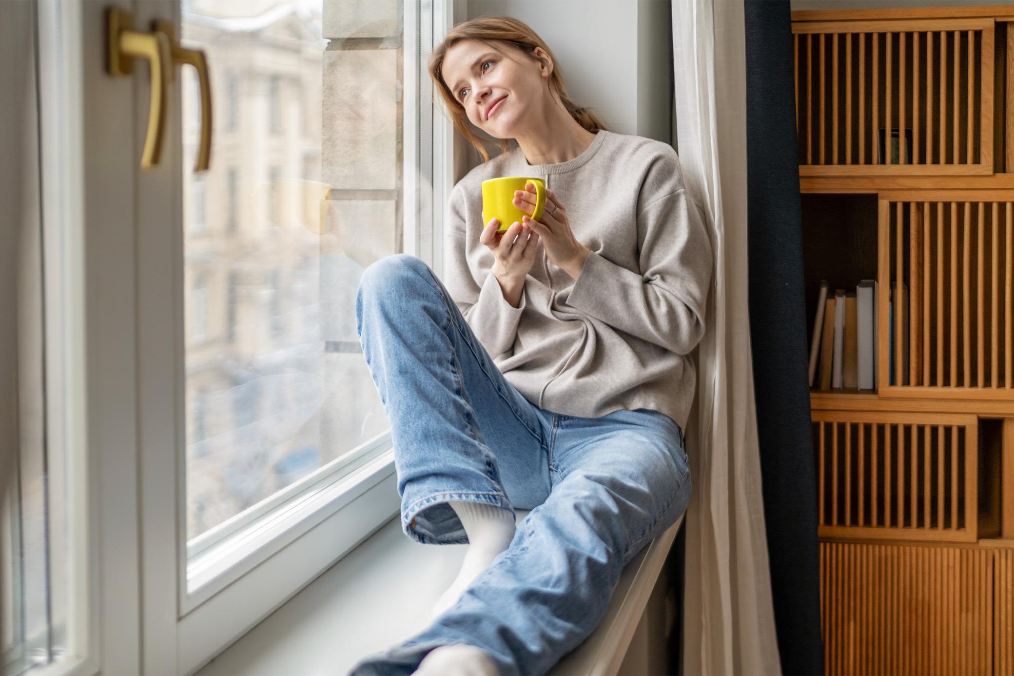 Immer up-to-date beim Gaspreis? Wir haben alle Infos. Frau sitzt auf Fensterbank über Heizung und hält eine gelbe Tasse in der Hand