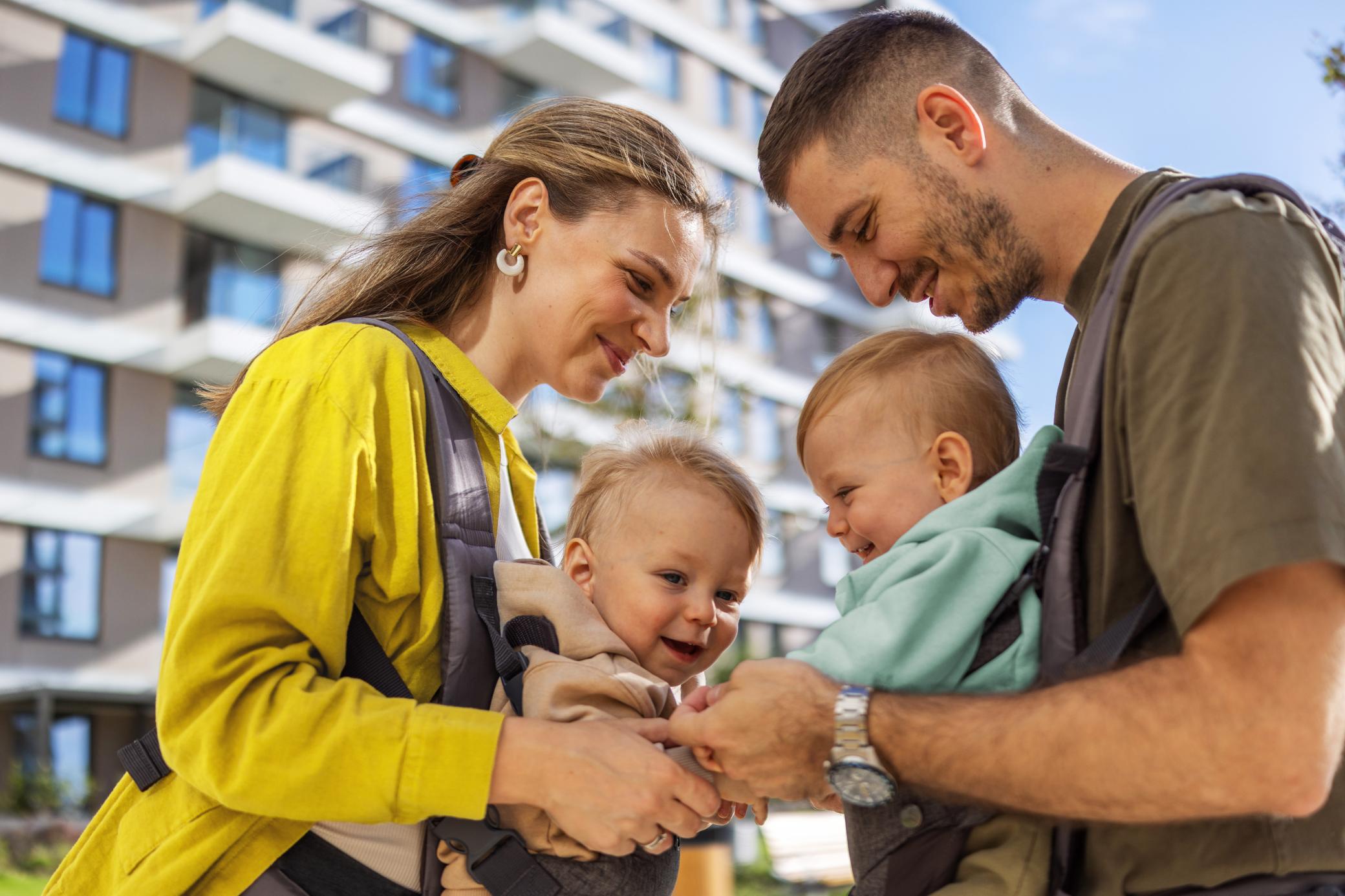 Ein glückliches Paar mit zwei Babys in der Trage steht vor einem Mehrfamilienhaus